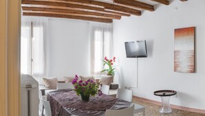 Un comedor con una mesa preparada para cuatro, una planta en maceta con flores, un televisor en la pared y un cuadro.