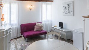 A small, cozy room with a purple sofa, a white table, a white cabinet, a television, and a washing machine. - Accademia Charm 5 by Wonderful Italy (Venecia)