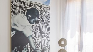 Een kamer met een groot abstract schilderij aan de muur, een kleine tafel met een plant en een speaker, en een raam met dunne gordijnen.