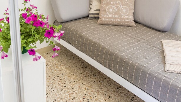 Een moderne woonkamer met een grijze bank, een decoratief kussen en een potplant met roze bloemen.