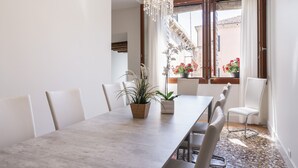 Un espace repas avec une longue table, des chaises, des plantes en pot et une vue sur un bâtiment par de grandes fenêtres ouvertes.