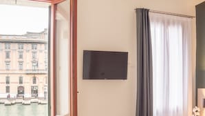 A hotel room with a bed, a TV mounted on the wall, a window with curtains, and a small wooden stool. - Camera 1 - Grand Canal Suites by Wonderful Italy (Venecia)