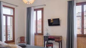 Habitación de hotel con cama, escritorio, silla, televisor y ventanas amplias con cortinas.