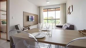 A modern dining room with a wooden table, chairs, a flat-screen TV mounted on the wall, and large windows offering a view of greenery outside. - Iloa Residence Resort Para 4 Pessoas by MME (Barra de São Miguel)