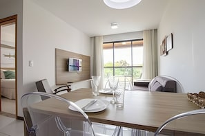 Comedor moderno con una mesa de madera, sillas, televisor de pantalla plana en la pared y ventanas amplias que dan a vistas de vegetación.