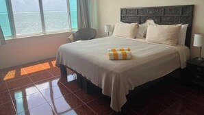 A hotel room with a large bed, a chair, a nightstand, and a window with a view of the sea. - Quiet Beach Front Condo with patio overlooking Infinity Pool & Ocean (Puerto Morelos)