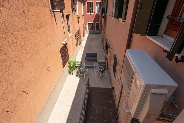Un callejón estrecho con un pequeño espacio para mesas al aire libre, un equipo de aire acondicionado grande y una puerta que da a un edificio.