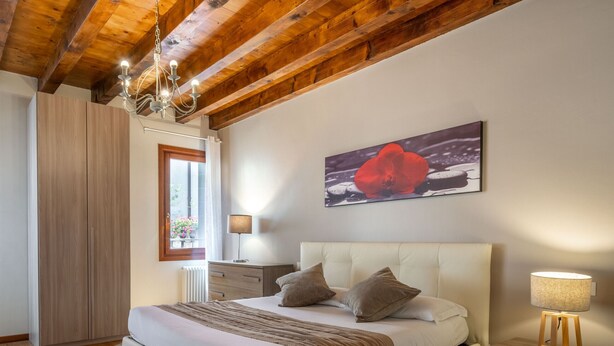 Un dormitorio con una cama grande, piso de madera, un candelabro, una mesita de noche con lámpara y una ventana con cortinas.