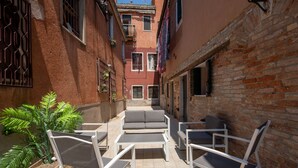 Un patio exterior estrecho con sillas de metal blanco y un sofá gris, rodeado de paredes de ladrillo y una planta en maceta.