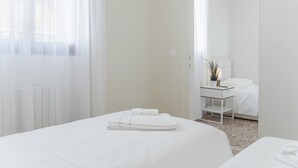 Ein schlicht eingerichtetes Hotelzimmer mit einem großen Bett, einem Nachttisch mit Lampe und Blick durch eine offene Tür.
