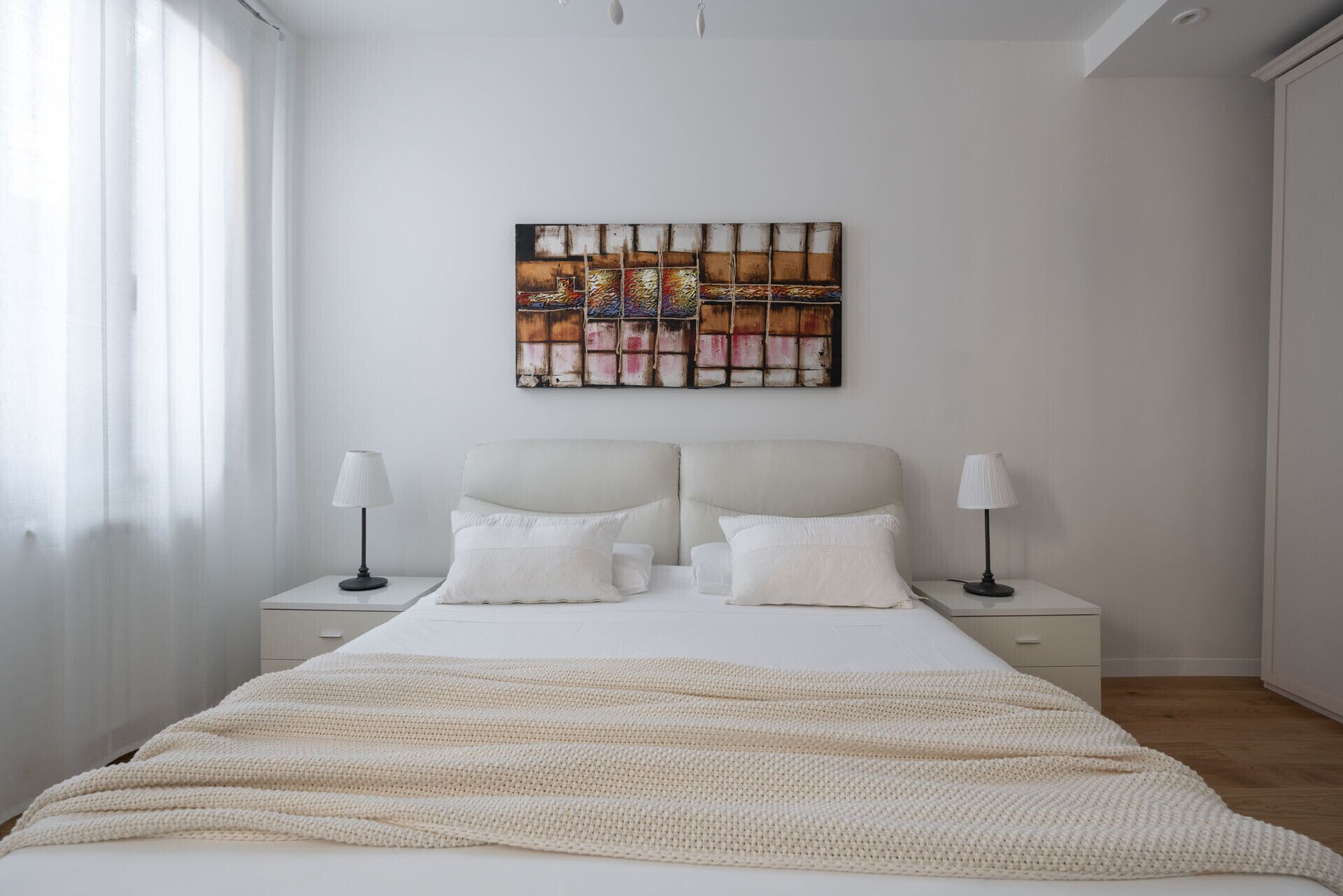 A bedroom with a large bed, two bedside tables with lamps, a white headboard, and a colorful abstract painting on the wall.