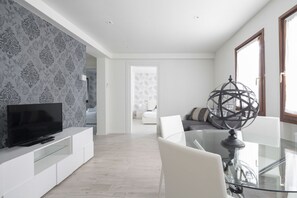 Un salon moderne avec un téléviseur à écran plat posé sur une console blanche, une table à manger en verre avec des chaises, et un globe décoratif sur un piédestal.