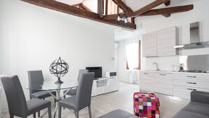 A modern kitchen with white cabinets and stainless steel appliances, a dining area with a glass table and gray chairs, and a colorful quilted ottoman. - San Marco Style Canal View Apt. 2 by Wonderful Italy (Venecia)