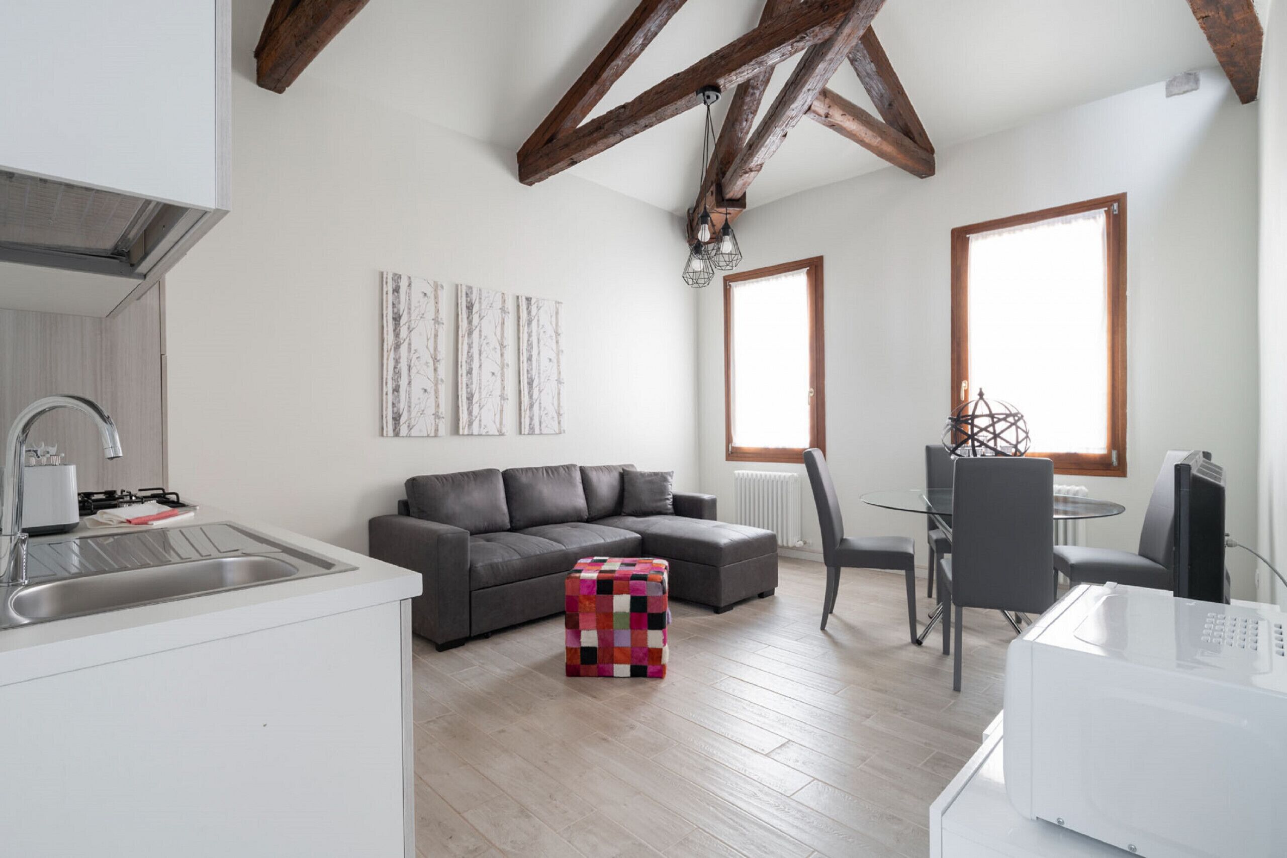 A modern kitchen with a built-in sink, a gray sofa, a colorful ottoman, and a dining area with a table and chairs.
