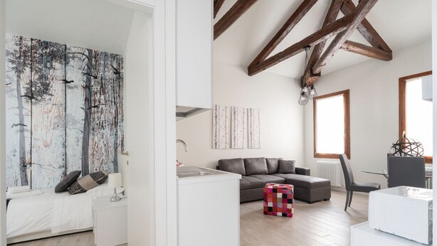 A modern living room with a sofa, a small table, and a colorful ottoman. There is a white kitchen area with a refrigerator and a sink. The room has wooden beams on the ceiling and large windows. - San Marco Style Canal View Apt. 2 by Wonderful Italy (Venecia)