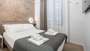 Une chambre d’hôtel avec un lit, deux serviettes roulées, une table de chevet, un téléviseur fixé au mur et une fenêtre avec des rideaux.