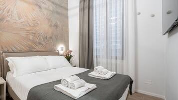 Ein Hotelzimmer mit einem Bett, zwei aufgerollten Handtüchern, einem Nachttisch, einem an der Wand befestigten Fernseher und einem Fenster mit Vorhängen.
