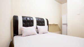 Una cama con cabecera capitonada, dos almohadas y una colcha blanca en una habitación con un armario blanco y una mesita de noche.