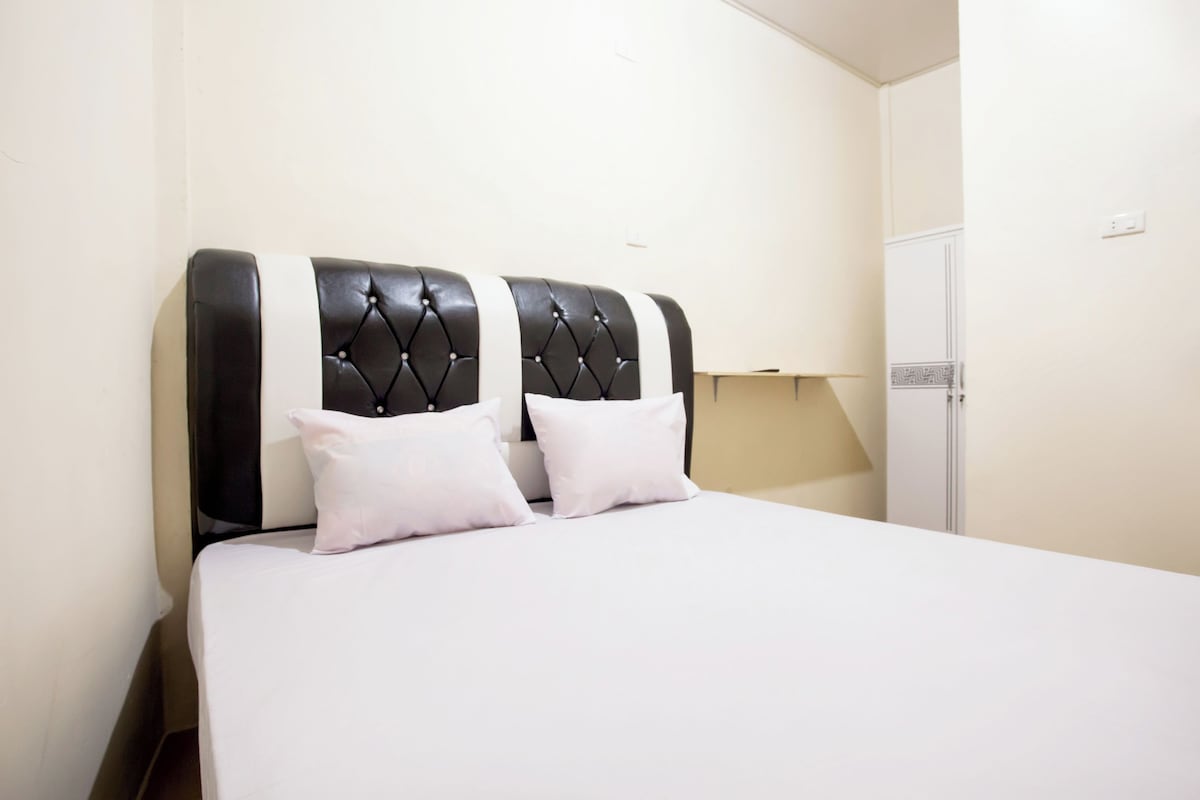 Una cama con cabecera capitonada, dos almohadas y una colcha blanca en una habitación con un armario blanco y una mesita de noche.