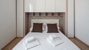 Ein ordentlich bezogenes Bett mit weißen Leinen und braunen Kissen, ein Holz-Kopfbrett und ein weißer Kleiderschrank darüber.