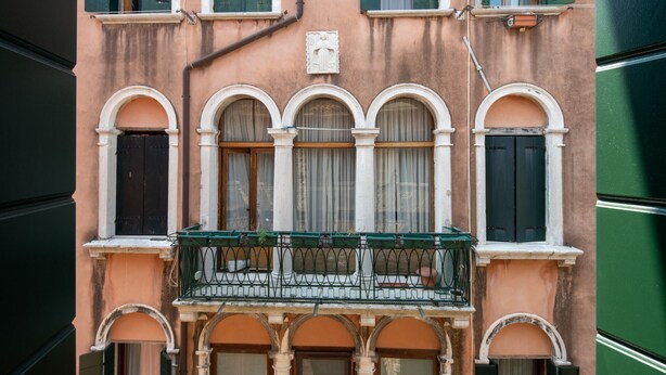 Ein Gebäude mit Rundbogenfenstern, einem Balkon mit Metallgeländer und einer kleinen Statue über den Fenstern.