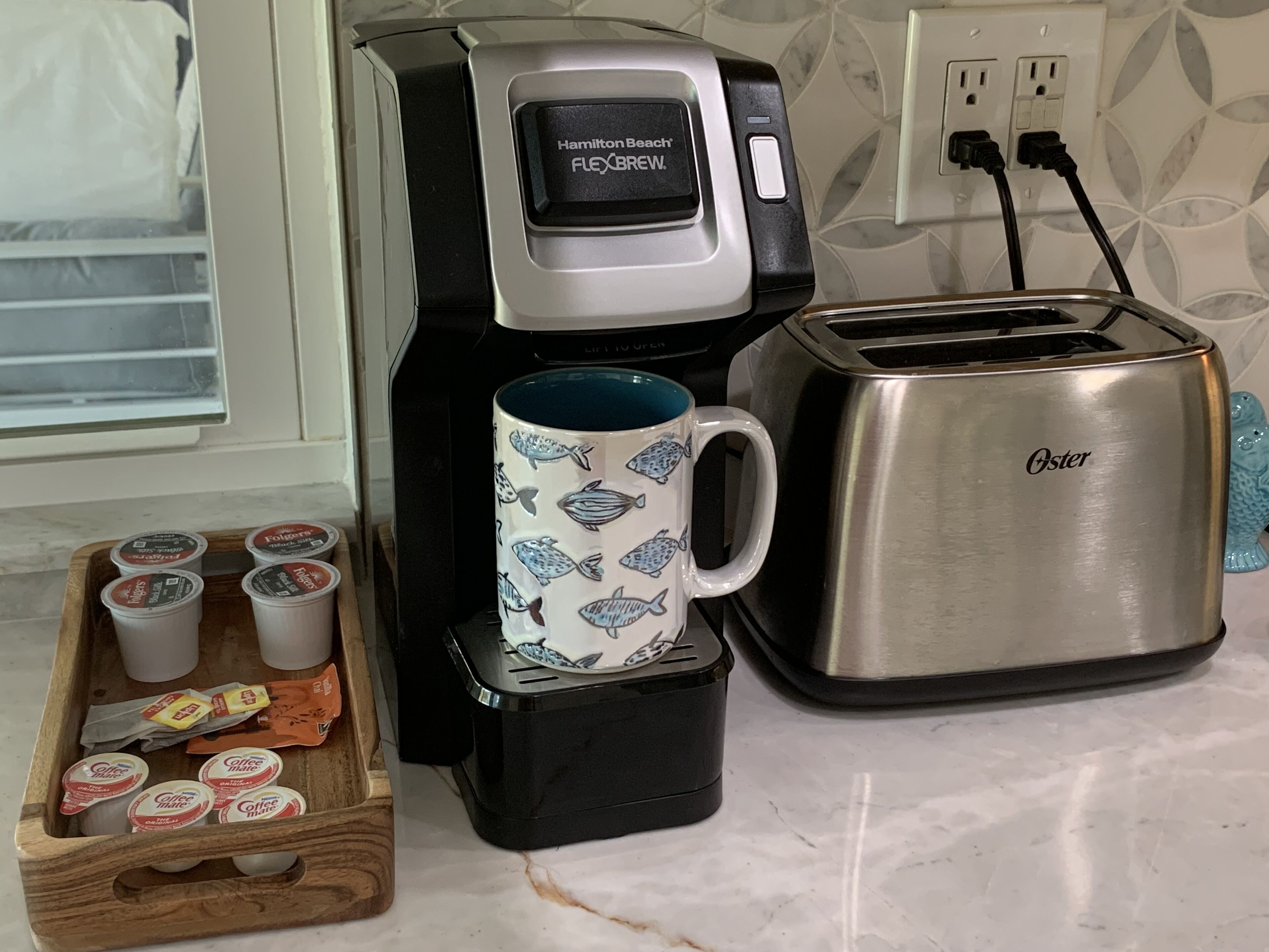 Una cocina con una cafetera Hamilton Beach Flexbrew, un tostador y una taza con diseños de peces. Hay cápsulas de café y una bandeja de madera con cápsulas de café y una bolsita de té.