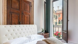 A bedroom with a white tufted headboard, a wooden wardrobe, a bedside table with a plant, and a window with a view of buildings and flowers. - Venice Luxury Palace 3 by Wonderful Italy (Venecia)