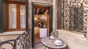 Un pequeño balcón con mesa y sillas, vista hacia una cocina y una reja ornamental de hierro forjado.
