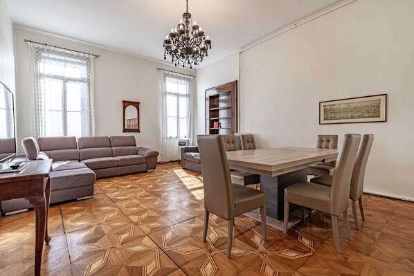 A living room with a sofa, a dining table with chairs, a chandelier, and a bookshelf.