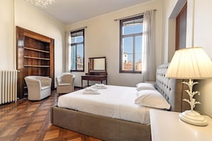 A hotel room with a large bed, two armchairs, a wooden wardrobe, a desk, and a lamp.