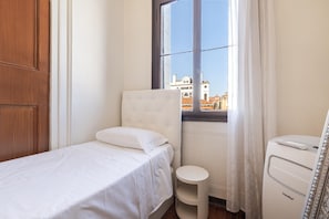 A small, neatly arranged bedroom with a single bed, a bedside table, a chair, and a window with a view of buildings.