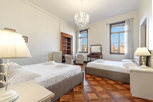 Quarto de hotel com duas camas, uma escrivaninha, uma cadeira, radiador e vista para a cidade pela janela.