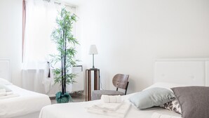 Habitación de hotel con dos camas, una planta grande en maceta, una lámpara y una silla.