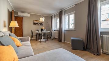 A modern living room with a grey sofa, a dining table with chairs, a radiator, and a window with curtains.