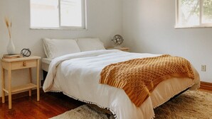 Une chambre à coucher comprenant un lit, deux tables de chevet, une petite table, un ventilateur fixé au mur et deux fenêtres.