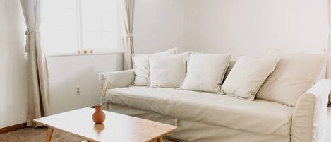 Un salón minimalista con un sofá color beige, una mesa de centro de madera y una ventana con cortinas.