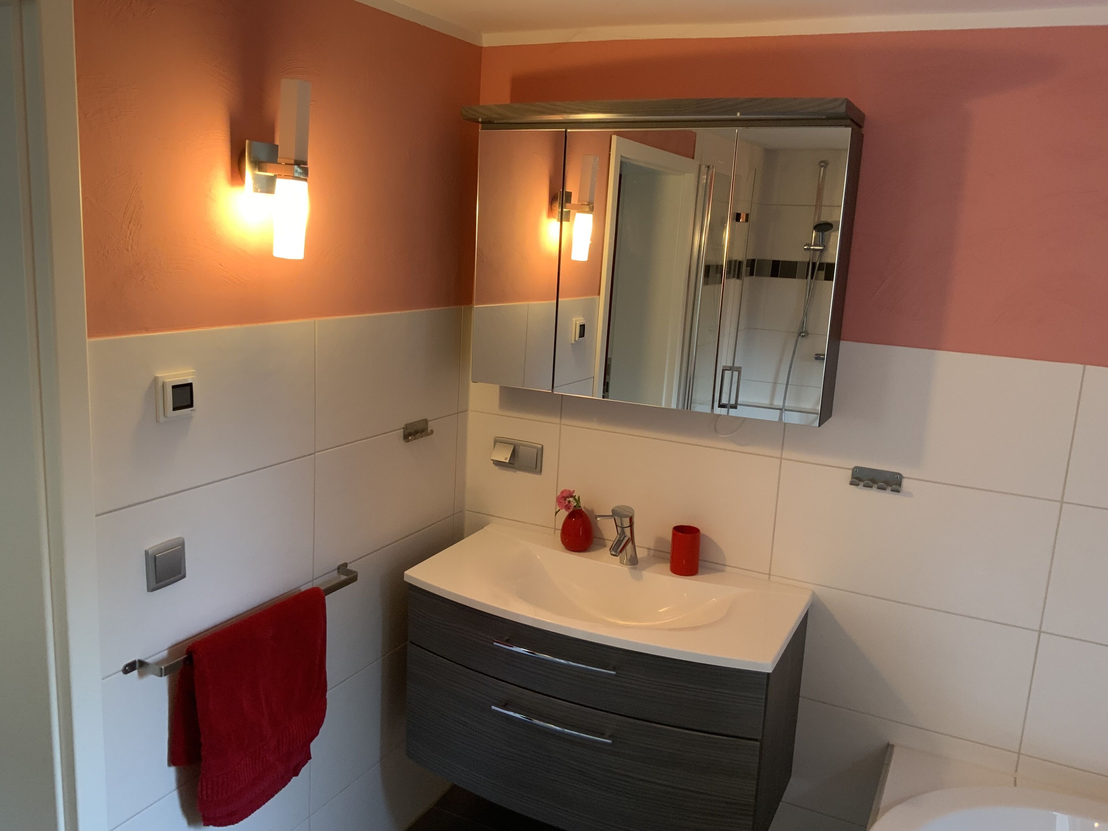 Un baño con un lavabo blanco, un espejo encima y una toalla roja colgada en un perchero.