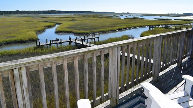 A wooden deck overlooking a marshy landscape with water channels and distant trees. - Fiddler on the Creek | Waterfront w/ Lighthouse Views, Kayaks & Dock (Chincoteague)