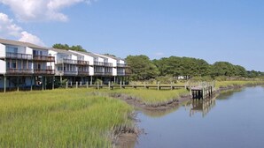 A waterfront building with multiple balconies, a dock, and a grassy area with a small water body. - Fiddler on the Creek | Waterfront w/ Lighthouse Views, Kayaks & Dock (Chincoteague)