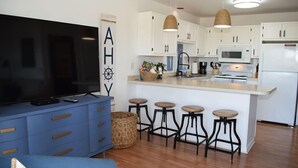 A modern kitchen with white cabinets, a central island with stools, a flat-screen TV, and a blue sofa. - Fiddler on the Creek | Waterfront w/ Lighthouse Views, Kayaks & Dock (Chincoteague)