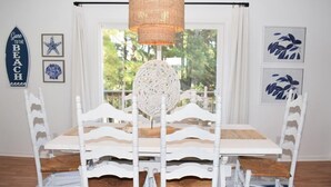 A dining area with a white table and chairs, a woven pendant light, and a view through a window. - Fiddler on the Creek | Waterfront w/ Lighthouse Views, Kayaks & Dock (Chincoteague)