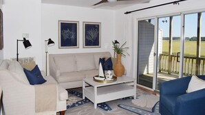 A living room with a sofa, coffee table, and a view through a sliding glass door to an outdoor deck. - Fiddler on the Creek | Waterfront w/ Lighthouse Views, Kayaks & Dock (Chincoteague)