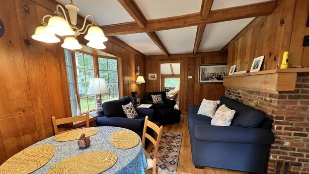 A cozy room with a wooden ceiling, a fireplace, a blue sofa, a dining table with placemats, and a window with a view of greenery. - 3BR/1BA Eastham cottage w/ backyard, A/C & WiFi (Eastham)