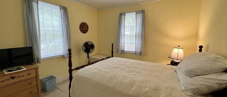 A bedroom with a bed, a bedside table, a lamp, a fan, and a television.