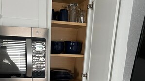 Ett kompakt kök med en mikrovågsugn, hyllor med burkar och skålar, ett torkställ för disk och en låda med köksredskap.