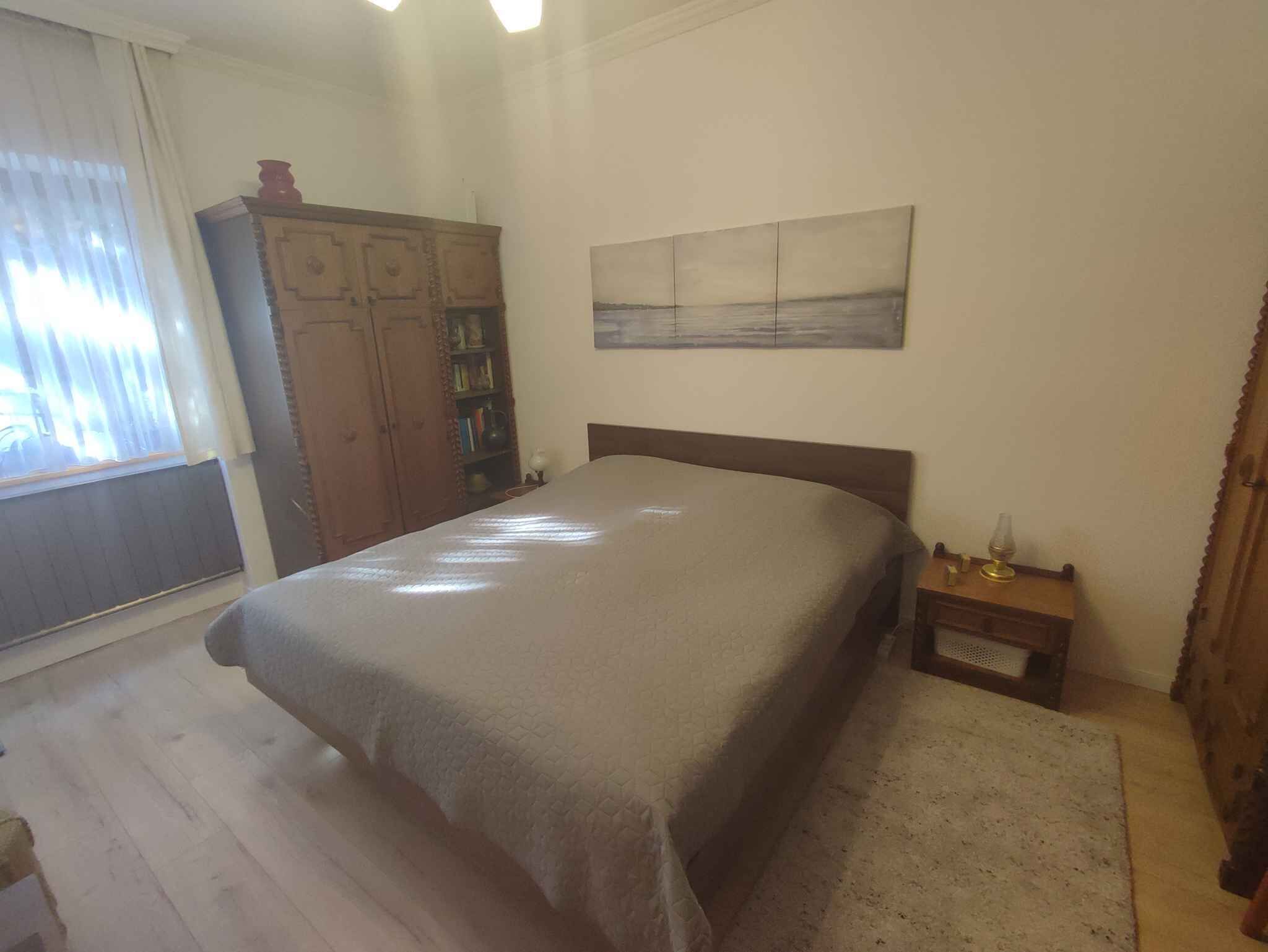 A bedroom with a bed, a wooden wardrobe, a nightstand, a lamp, and a painting on the wall.