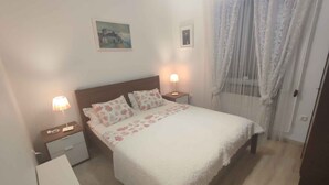A neatly made bed with floral bedding, two bedside lamps, a small bedside table, and a window with sheer curtains. - Holiday home with wifi and air conditioning (Balatonboglár)