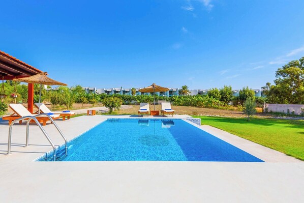 A swimming pool with lounge chairs and umbrellas, surrounded by greenery and buildings in the background.