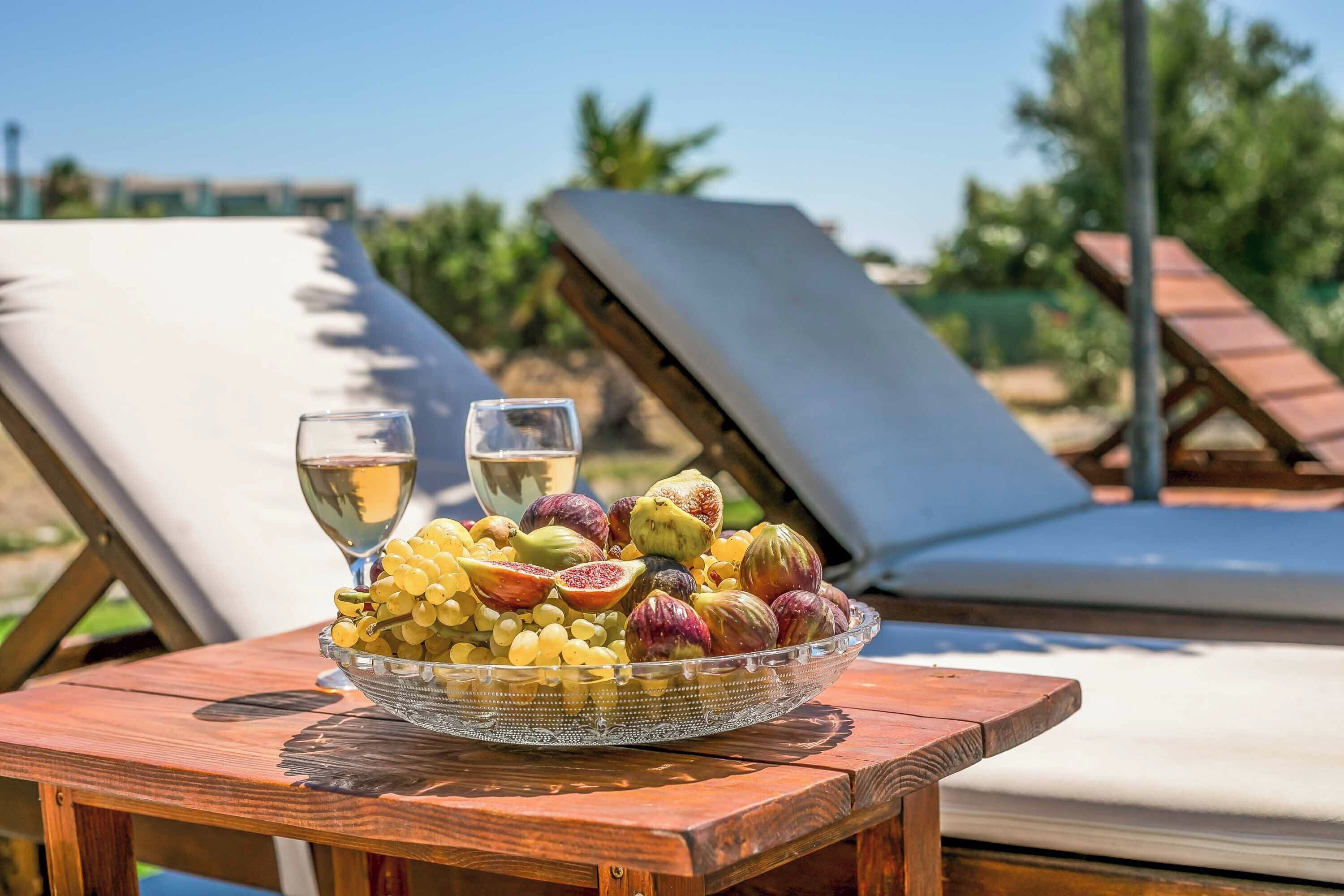 A wooden table with a glass bowl of fruits and two glasses of white wine, set against a backdrop of sun loungers and greenery.