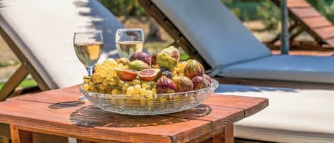 A wooden table with a glass bowl of fruits and two glasses of white wine, set against a backdrop of sun loungers and greenery.